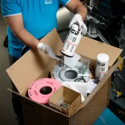 A person in white gloves and a blue shirt unpacks an Atlas Copco ZT15-22 Inlet Valve Overhaul Kit (P/N 2901110200), revealing filters, mechanical parts, a pink ring, metal springs, and sealed containers from the cardboard box.