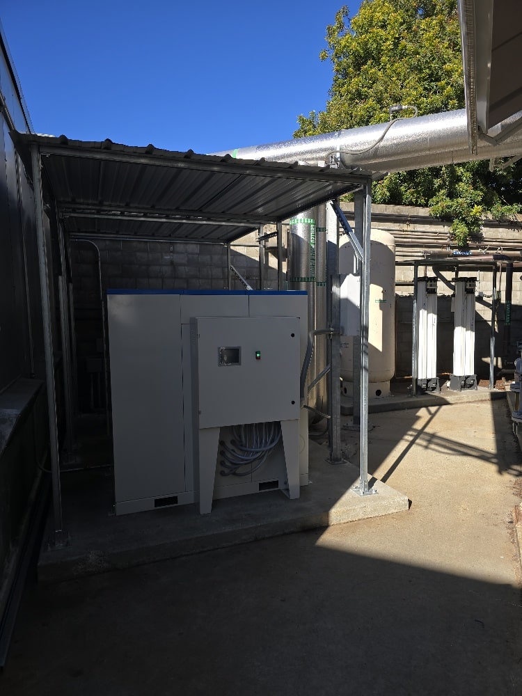 A white industrial machine sits under a metal canopy next to large pipes and storage tanks outdoors, with sunlight casting shadows on the concrete ground. Trees and a clear blue sky are visible in the background.