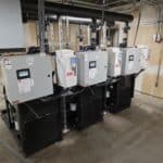 Four large industrial electrical control panels are mounted on black units in a utility room, with pipes, cables, and conduits running overhead and along the walls. The floor is concrete and the walls are unfinished.