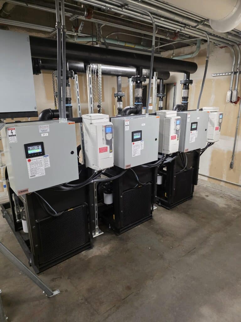 Four large industrial electrical control panels are mounted on black units in a utility room, with pipes, cables, and conduits running overhead and along the walls. The floor is concrete and the walls are unfinished.