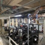 Industrial mechanical room with multiple large black pumps and pipes, control panels, and overhead exposed ceiling with various conduits and insulation. Safety rails and labeled equipment are visible throughout the area.