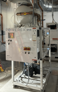 A Powerex, Inc. Laboratory Vacuum System (P/N LVPDT0502-DD-TM-AES-SPL) is installed in a utility room with visible cables, pipes, switches, and warning labels; a second identical unit stands behind it.