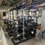 Three large black industrial pumps with exposed piping and wiring installed in a utility room with concrete floors, visible ceiling pipes, and various electrical panels on the walls.