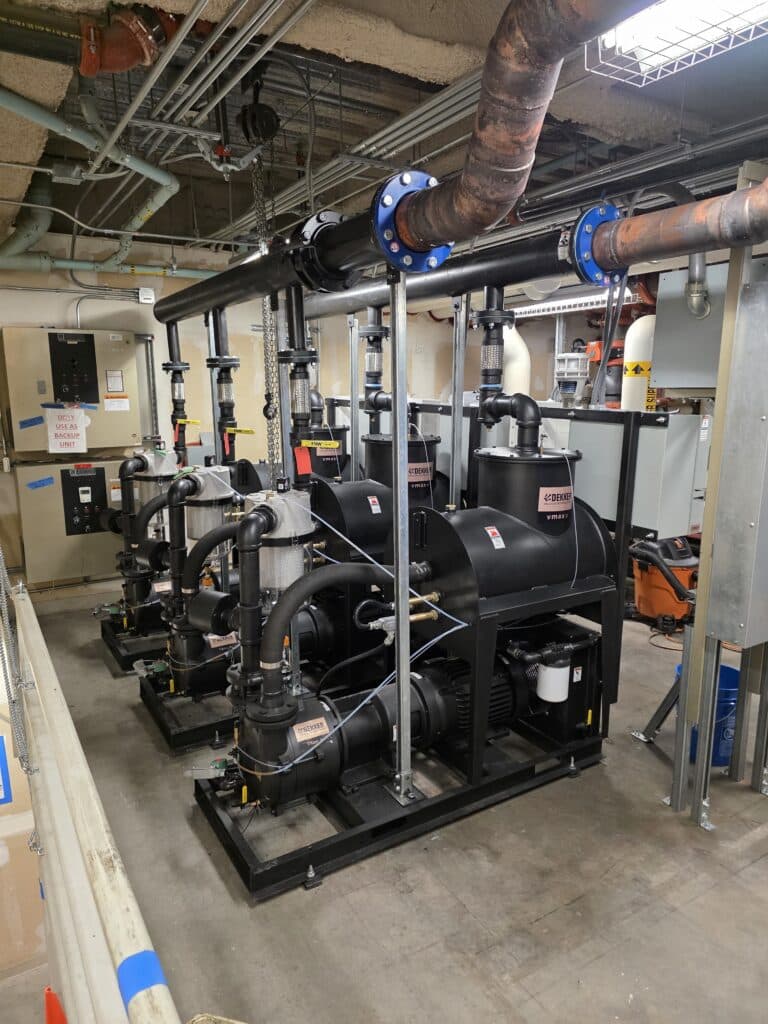 Three large black industrial pumps with exposed piping and wiring installed in a utility room with concrete floors, visible ceiling pipes, and various electrical panels on the walls.