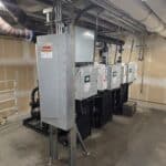 Industrial electrical panels and equipment are installed in a utility room with exposed pipes, conduit, and unfinished walls. Warning labels are visible on the panels, and the concrete floor is clear.