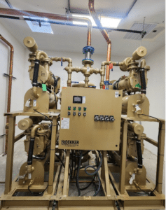 A Dekker Laboratory Vacuum System Model VMX0153MA4 40HP (P/N VMX0153MA4-AES-SPL) with multiple pipes and control panel is installed in a utility room with exposed copper piping and concrete flooring.