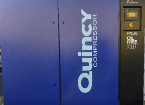 The Quincy Compressor QOFT-50V Oil-Free Compressor (P/N QOFT-50V-AES-SPL) is a large blue industrial unit with a right-side control panel and display, labeled "Quincy Compressor" and "OIL FREE" on the front.