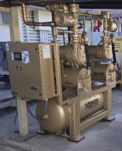 Dekker Laboratory Vacuum System Model VMX0153MA2-AES-SPL- Tank Mount (P/N VMX0153MA4-AES-SPL) with metal pipes, gauges, and a control box, installed outdoors on a concrete base. The equipment appears well-maintained and is connected to multiple utility lines.