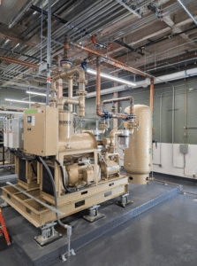Dekker Laboratory Vacuum System Model VMXVFD0553MA2-AES-SPL with pipes and control panels in a clean, modern mechanical room, surrounded by exposed ceiling pipes, grey flooring, and a large cylindrical tank beside the equipment.