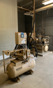 Two beige Dekker Laboratory Vacuum System units, Model VMX0036MA1-AES-SPL (P/N VMX0036MA1-AES-SPL), each equipped with an attached control panel, are positioned on a concrete floor in a utility room with exposed pipes and overhead lighting.