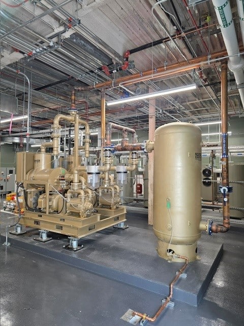 Industrial room with exposed ceiling pipes, large tan machinery including a vertical tank and pumps, on a gray floor. Electrical panels and additional piping are visible in the background. Bright lighting illuminates the space.