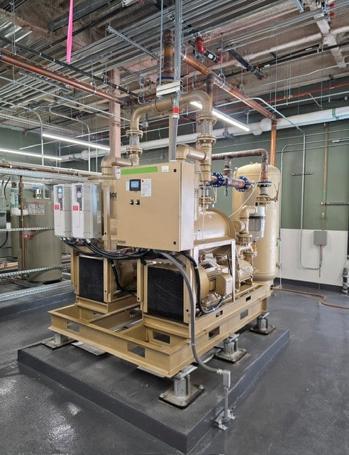 Large industrial machinery with beige metal pipes and equipment, mounted on a platform in a facility with exposed ceiling pipes, electrical panels, and polished concrete floors.