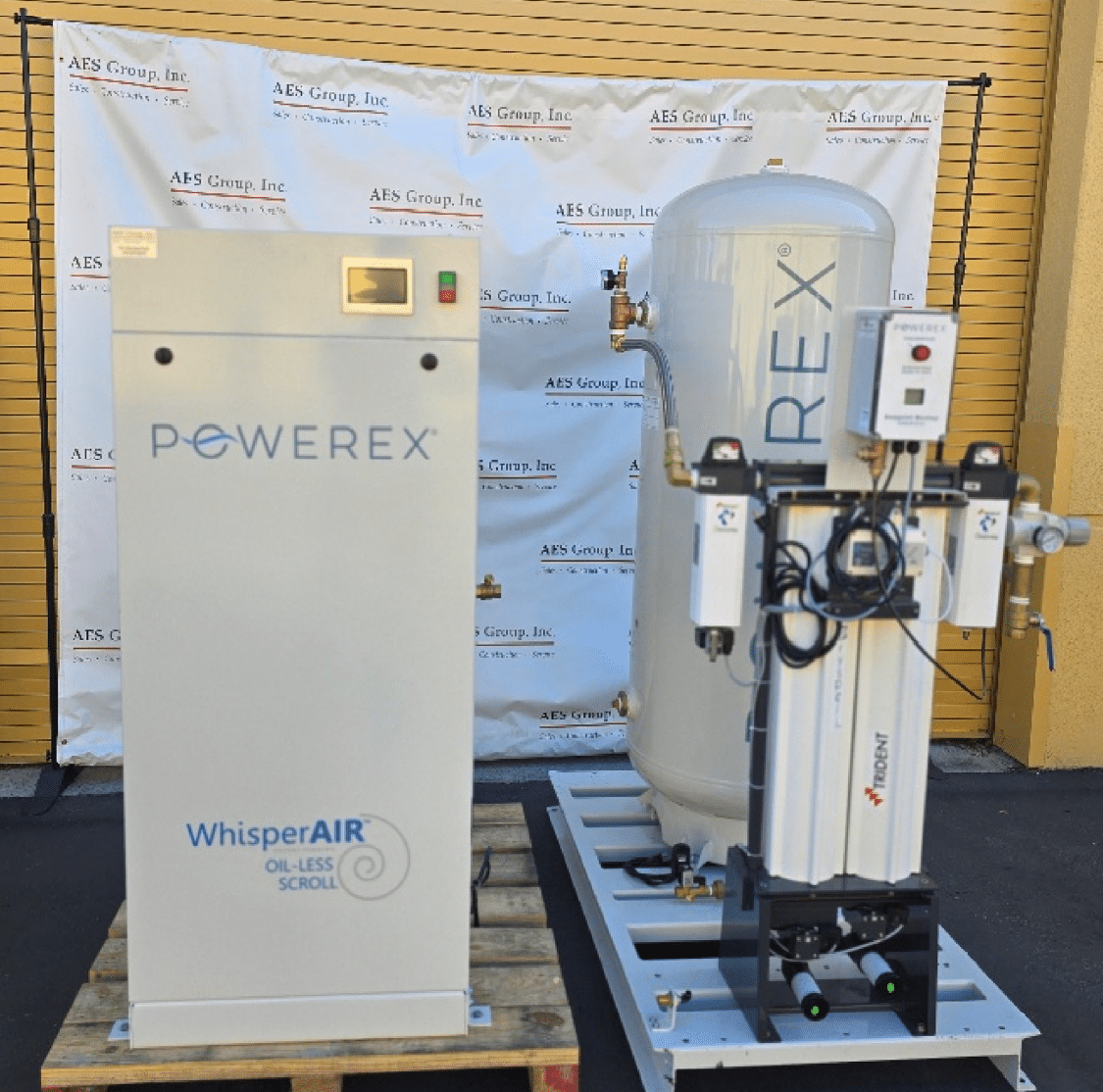 Two large industrial machines, including a Powerex Laboratory Compressed Air System Model SED1007-DD-AES-SPL (P/N SED1007-DD-AES-SPL) and a unit labeled “WhisperAIR,” are positioned outdoors on a blacktop, with an AES Group, Inc. branded backdrop and a yellow roll-up door in the background.