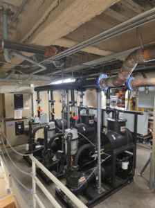A mechanical room with several large black pumps, pipes, valves, and electrical panels under an exposed concrete ceiling. Overhead, various conduits and wires—alongside a Dekker Laboratory Vacuum System Model VMXVFD0303KA3-AES-SPL unit—are visible.