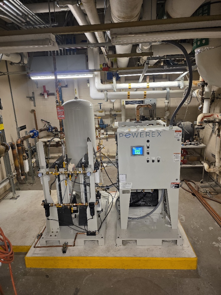 A Powerex medical air compressor system with a vertical receiver tank, control panel, and connected plumbing, installed on a concrete base in a mechanical room.