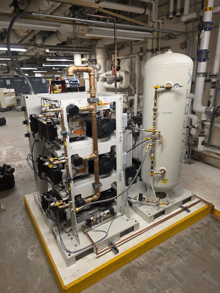 An industrial air compressor system featuring a vertical white storage tank and multiple pump units connected by copper piping, mounted on a yellow-edged concrete platform in a utility room.